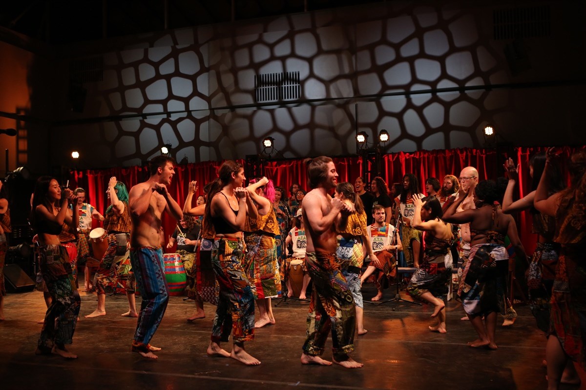 A group of performers dancing energetically on stage in colorful, patterned costumes, with musicians playing drums in the background.