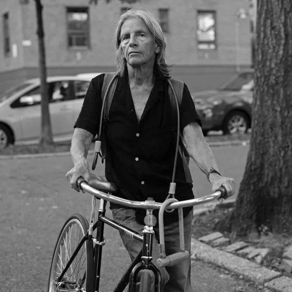 Eileen Myles, standing holding a bike, shot in black and white. 