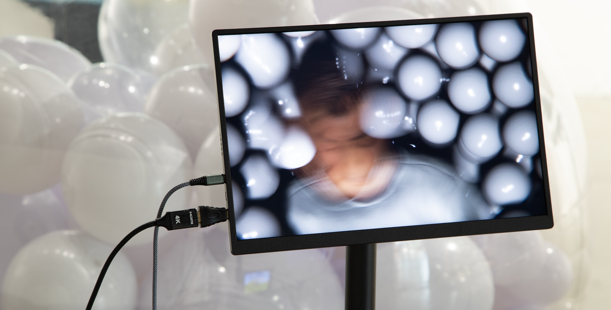 A monitor displays a blurred head amid a field of white balloons. Many white balloons are grouped behind the monitor.