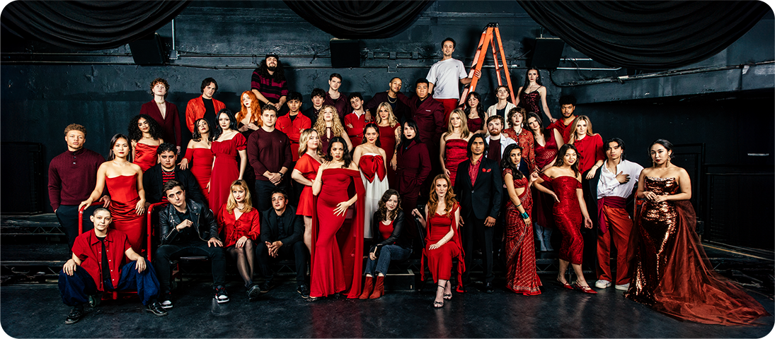 A large group of people wearing predominantly red and black formal attire, posed in a theatrical setting with black draped fabric in the background.