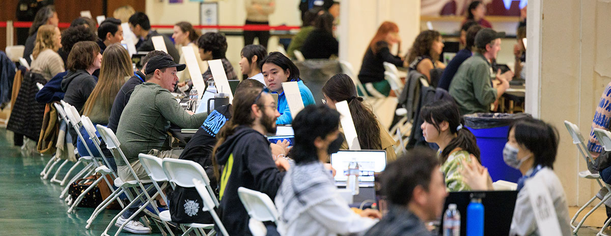 People seated at long tables in a busy indoor event, working on laptops and engaging in discussions.