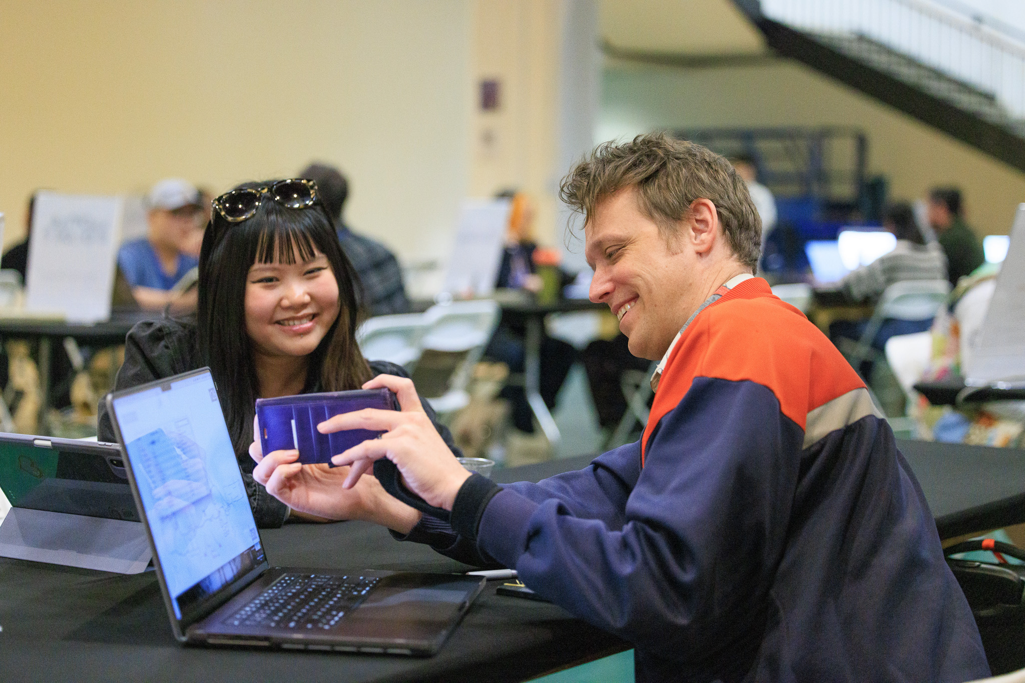 Two people at a table smiling and interacting, with a laptop and phone in front of them.