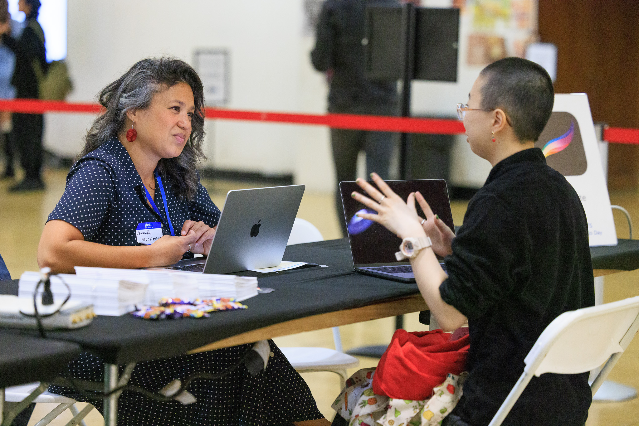 Two people seated at a table, talking, with laptops open.