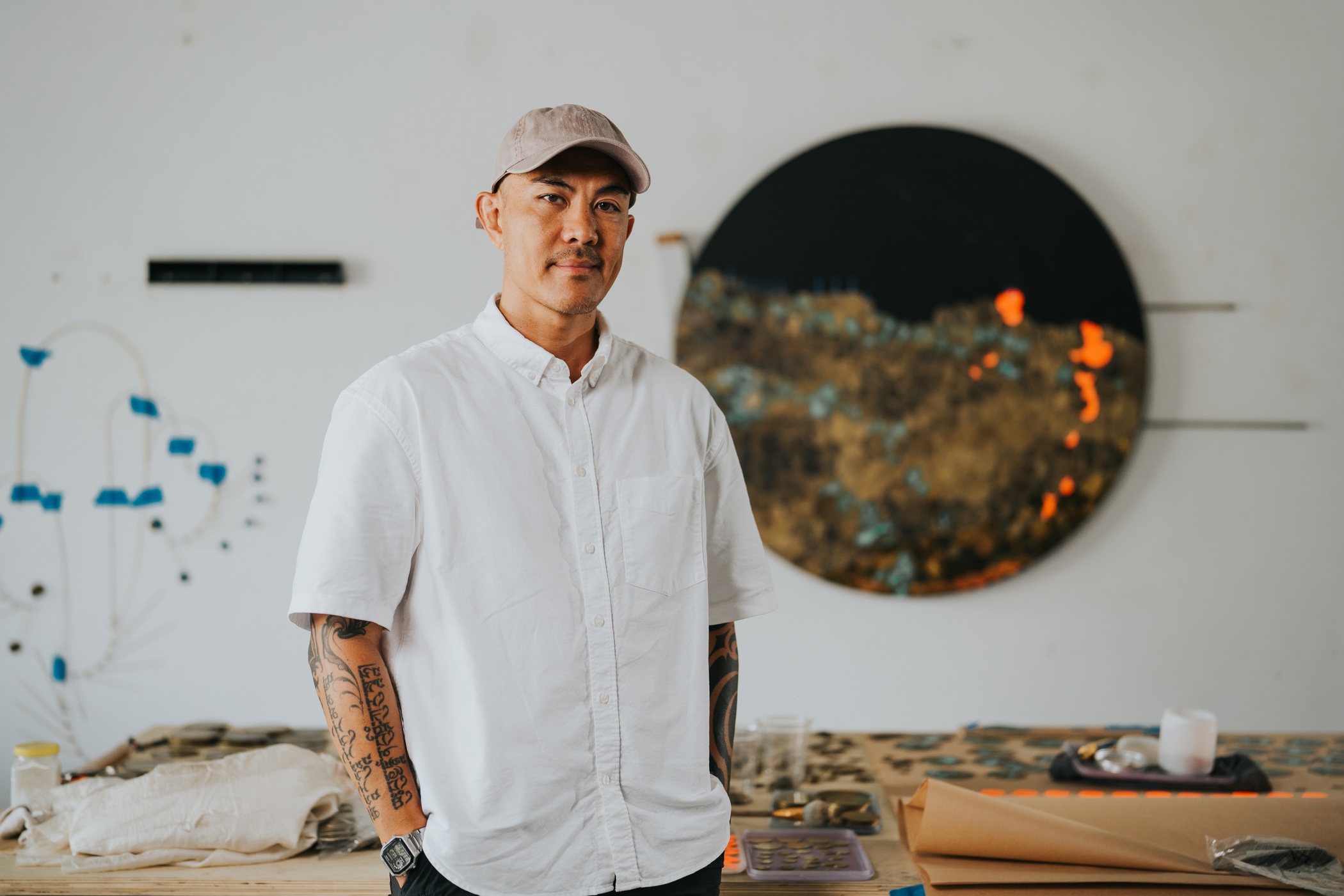 Tuan Andrew Nguyen, Multidisciplinary Artist, wearing a short-sleeved white shirt and baseball cap standing in front of a wall and table displaying his artworks. 