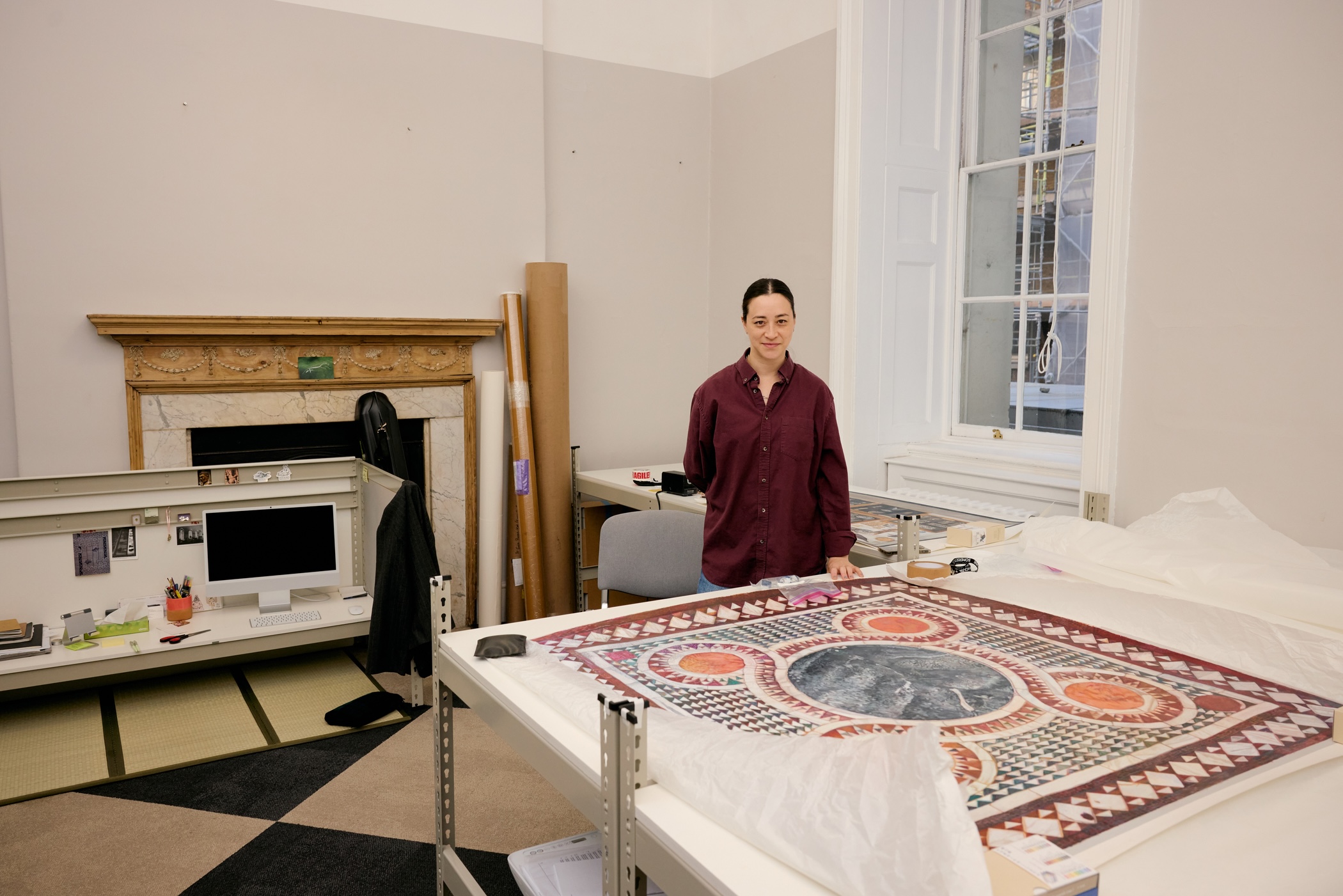 Gala Porras-Kim, Interdisciplinary Artist, wearing a burgundy shirt standing behind a table displaying her artwork in a studio. 