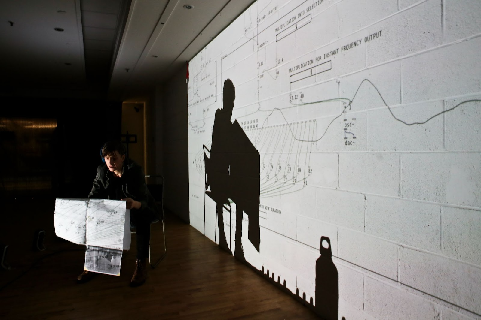 A person holds a paper in a dark room with technical drawings projected on a wall.