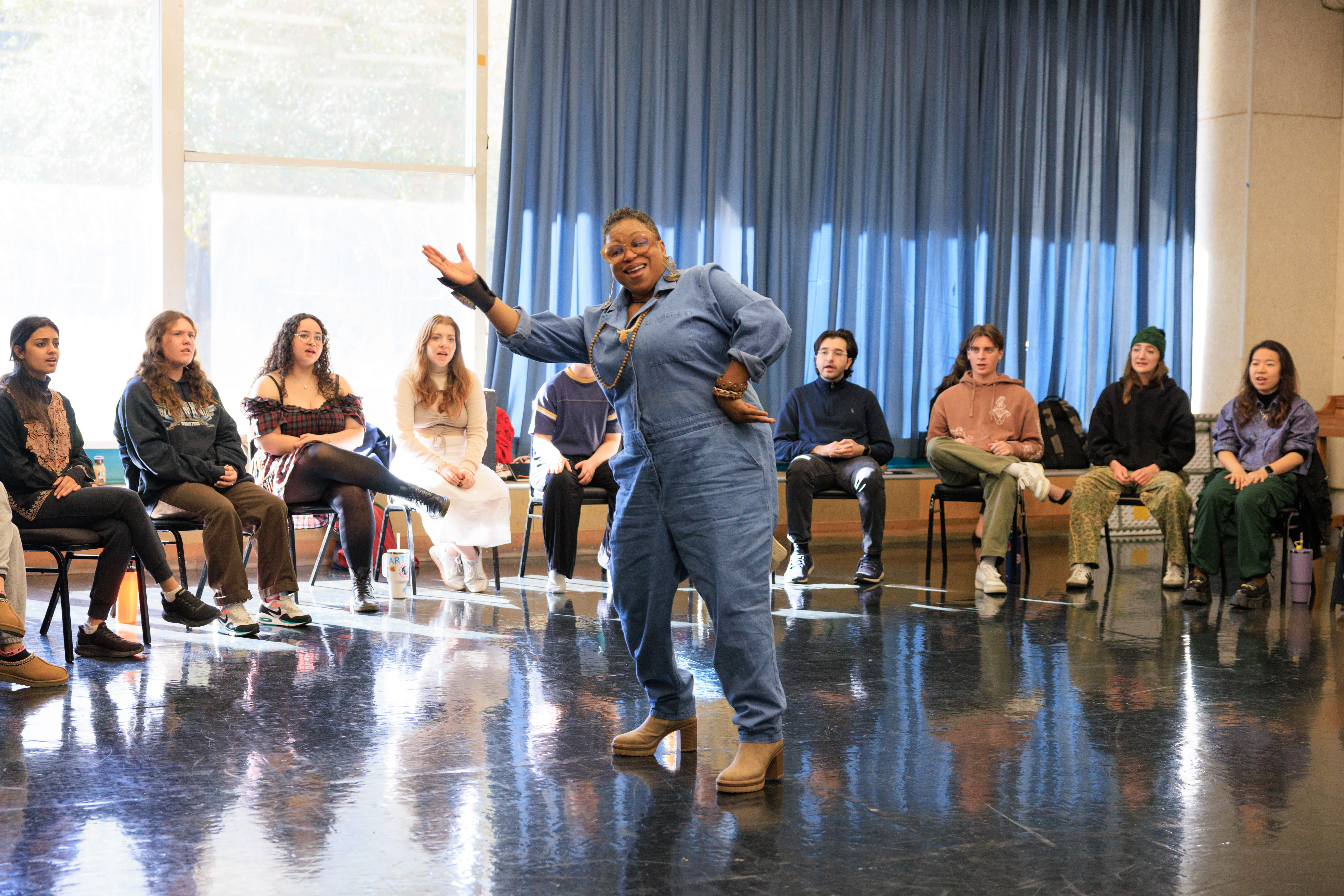 A person in a denim jumpsuit stands and gestures in front of a seated group in a room with large windows and blue curtains.