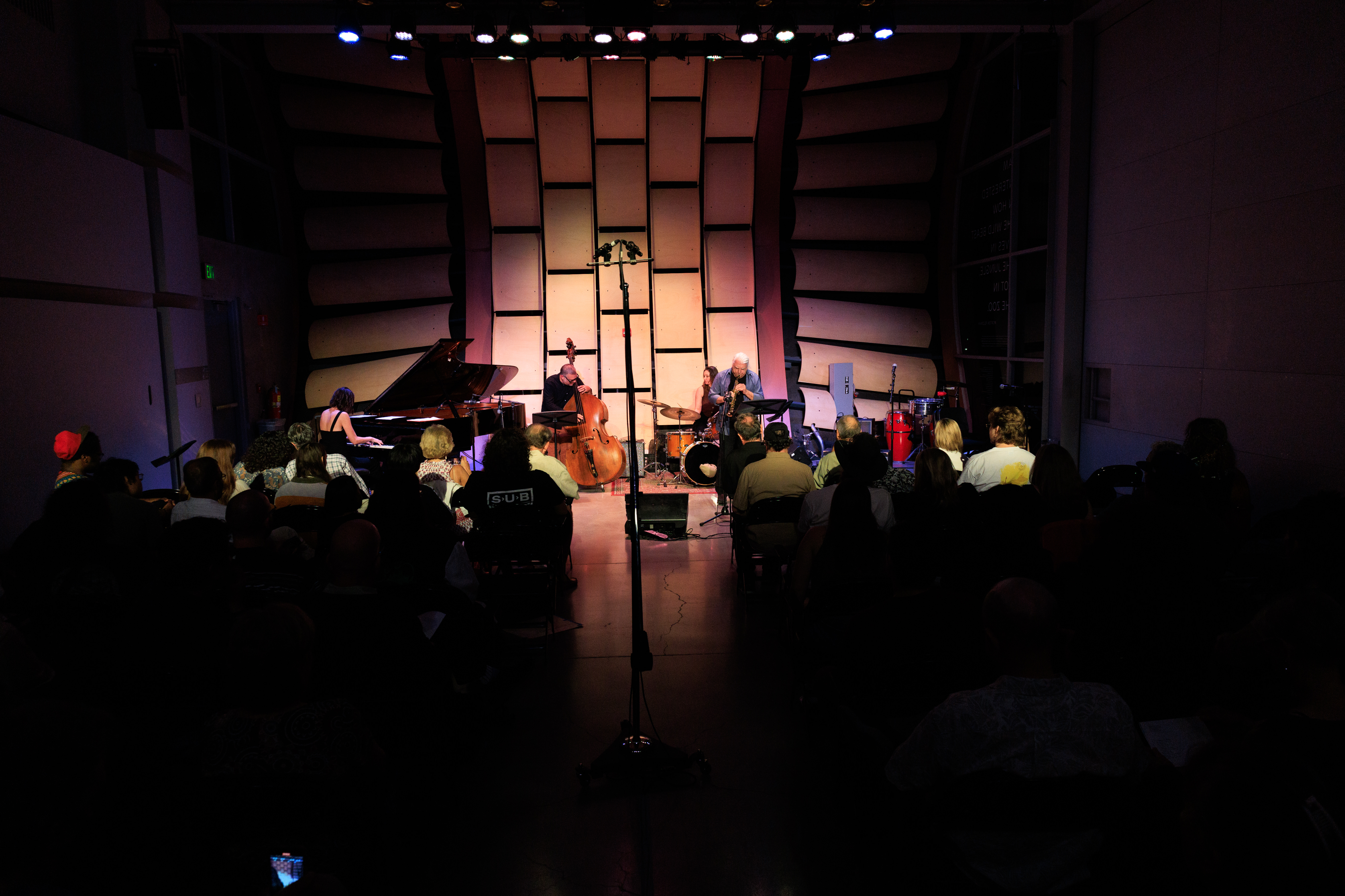 Jazz trio performing on a stage with a pianist, bassist, and drummer, viewed by an audience.