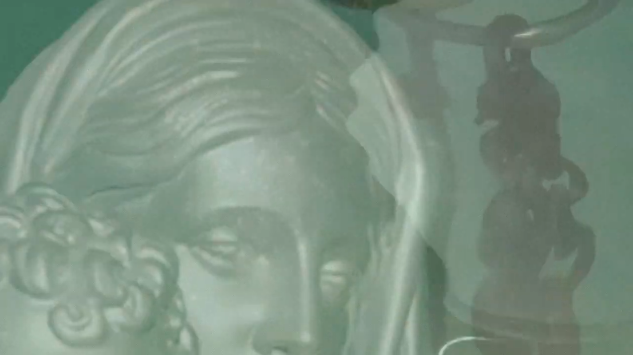Close-up of a sculpted face with a green hue and a shadowy figure in the background.