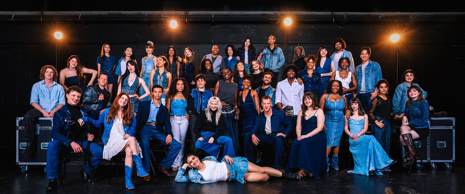 A diverse group of people in denim clothing posed on a stage with overhead lights.