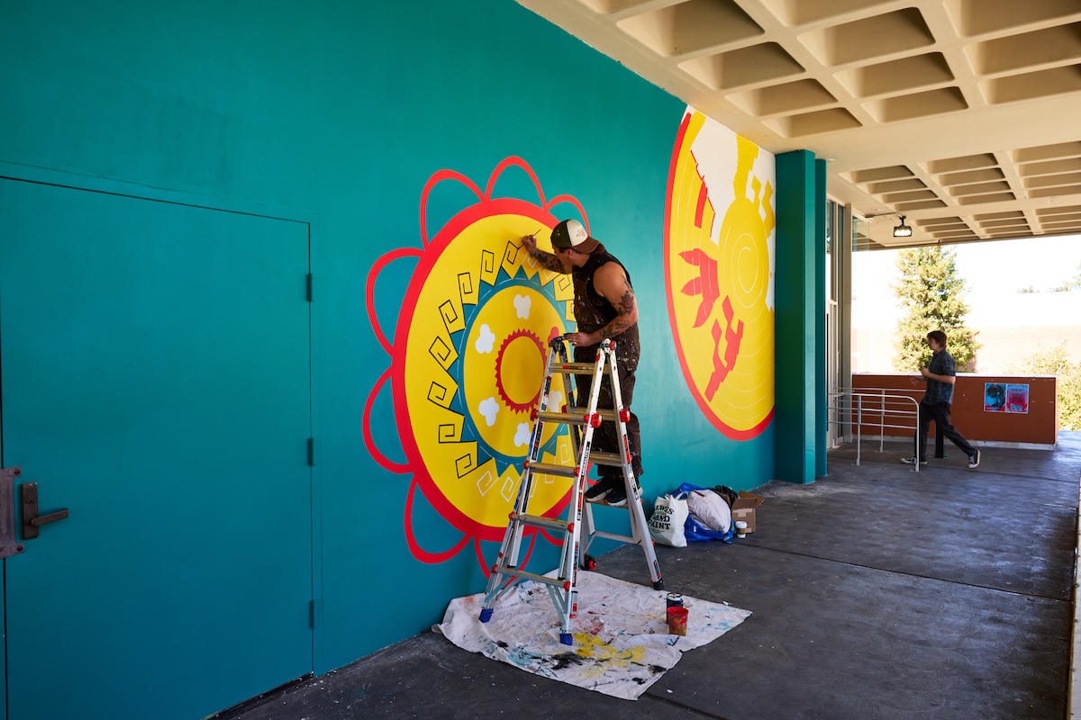 A man stands on a ladder while painting a sunlike mural on an aqua colored wall. 