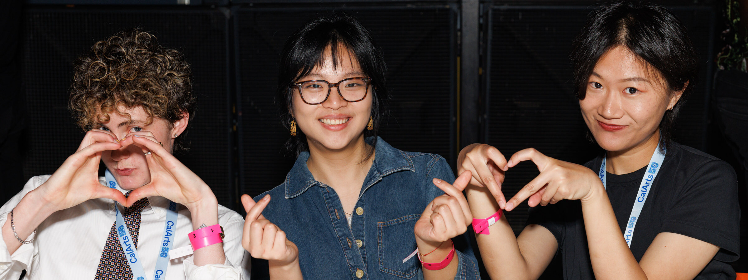 Three CalArts students make different heart shapes with their hands, smiling at the camera