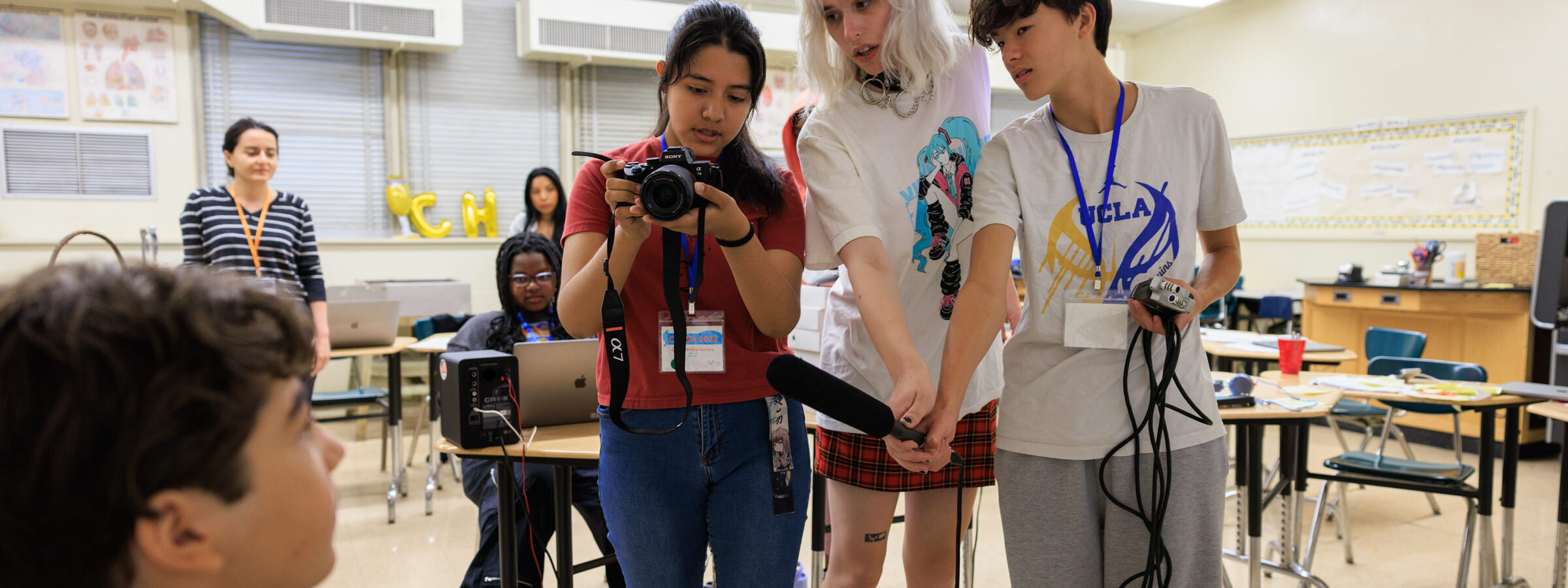 A group of three high school students look at a small camera screen while adjusting a microphone. 