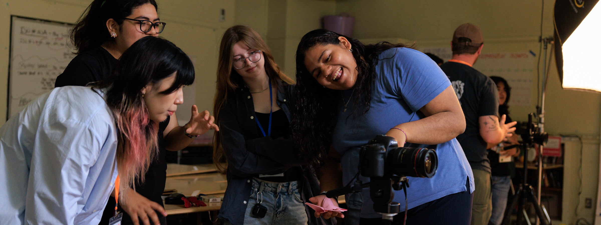 A group of four high school students examine a screen on a camera, laughing together. 