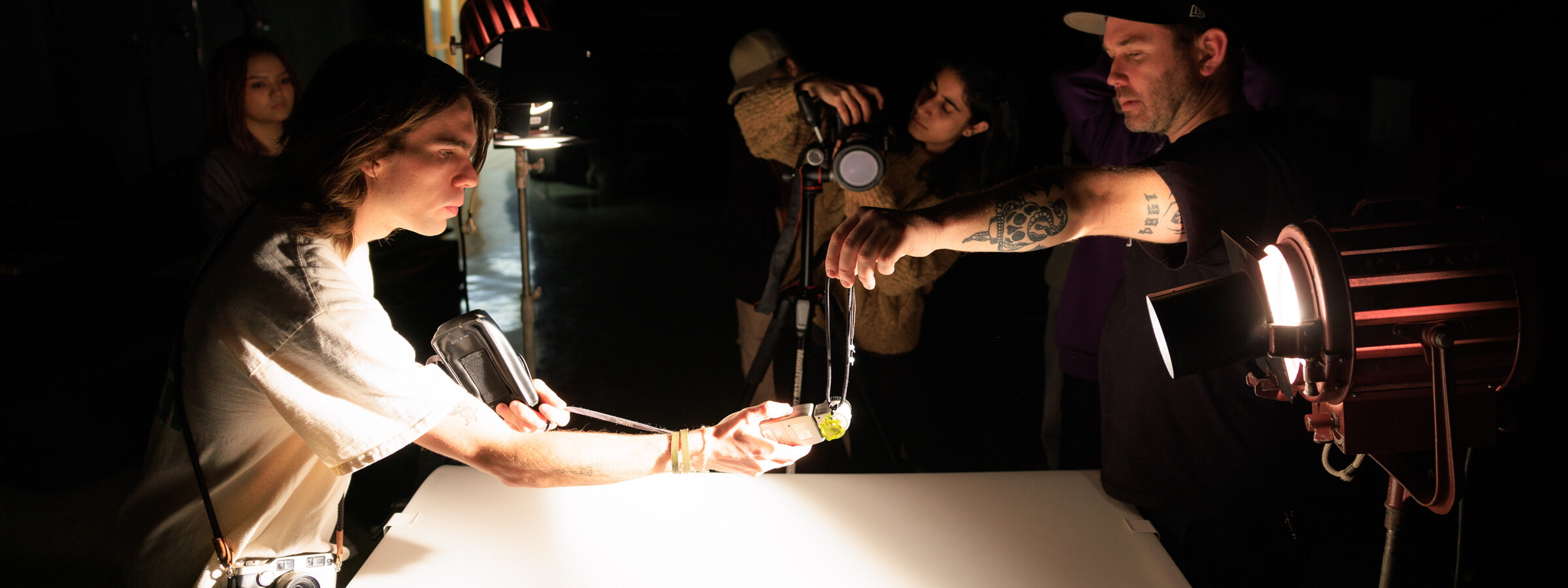 A group of four people in a dark room point photo lights and cameras at an object they’re holding above a dramatically lit white table. 