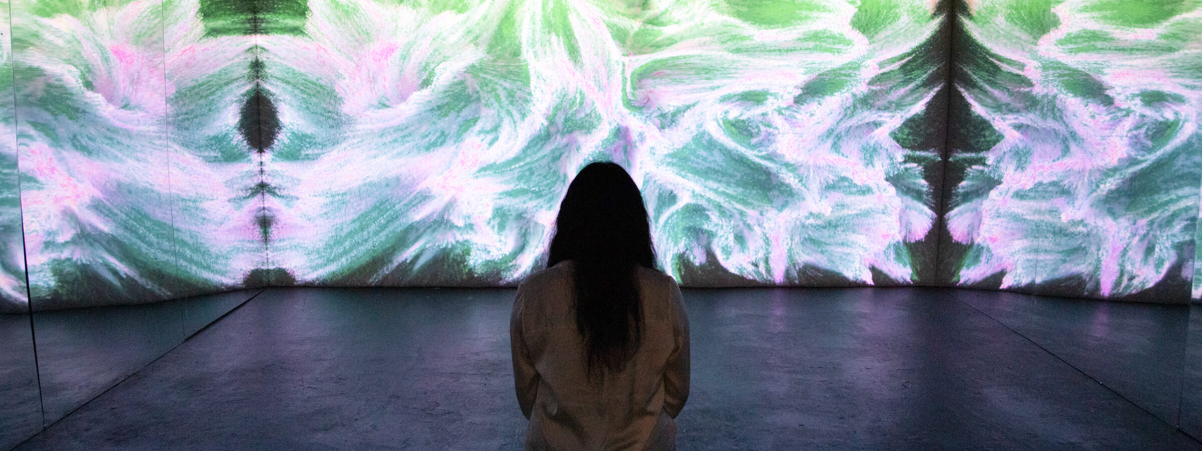 A person with long dark hair with their back to the camera faces a wall in a darkened art gallery. A fluid, abstract pattern in colors of pink, white, and green is projected on the wall.