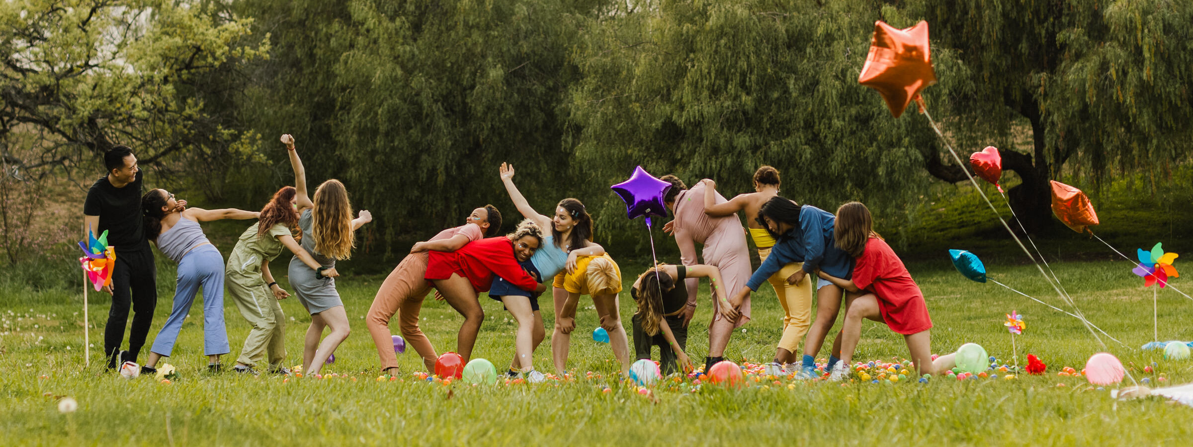 A line of 13 dancers in a grassy field twist their bodies, reaching for each other, with bright mylar balloons.