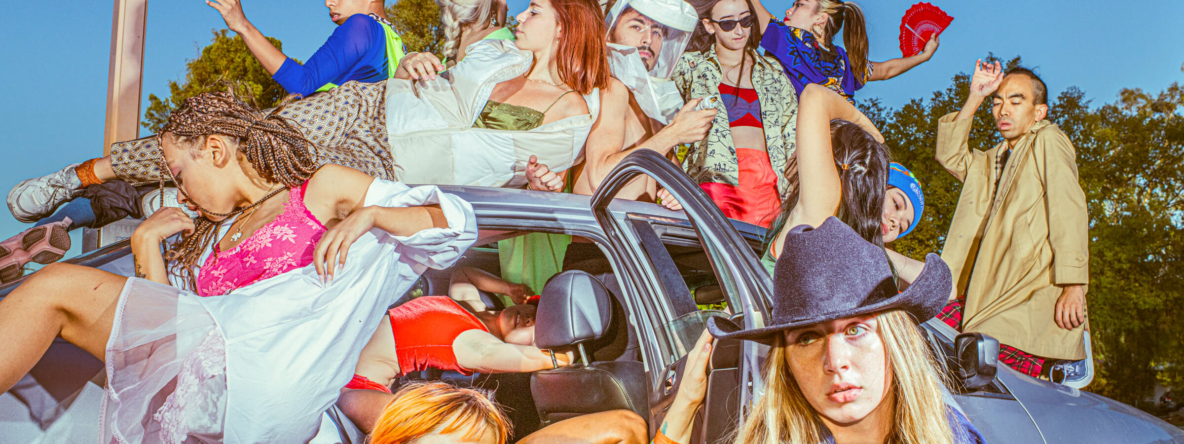 A large group of dancers in bright costumes, draped inside and across the top of a car. A woman in a cowboy hat is in the foreground.