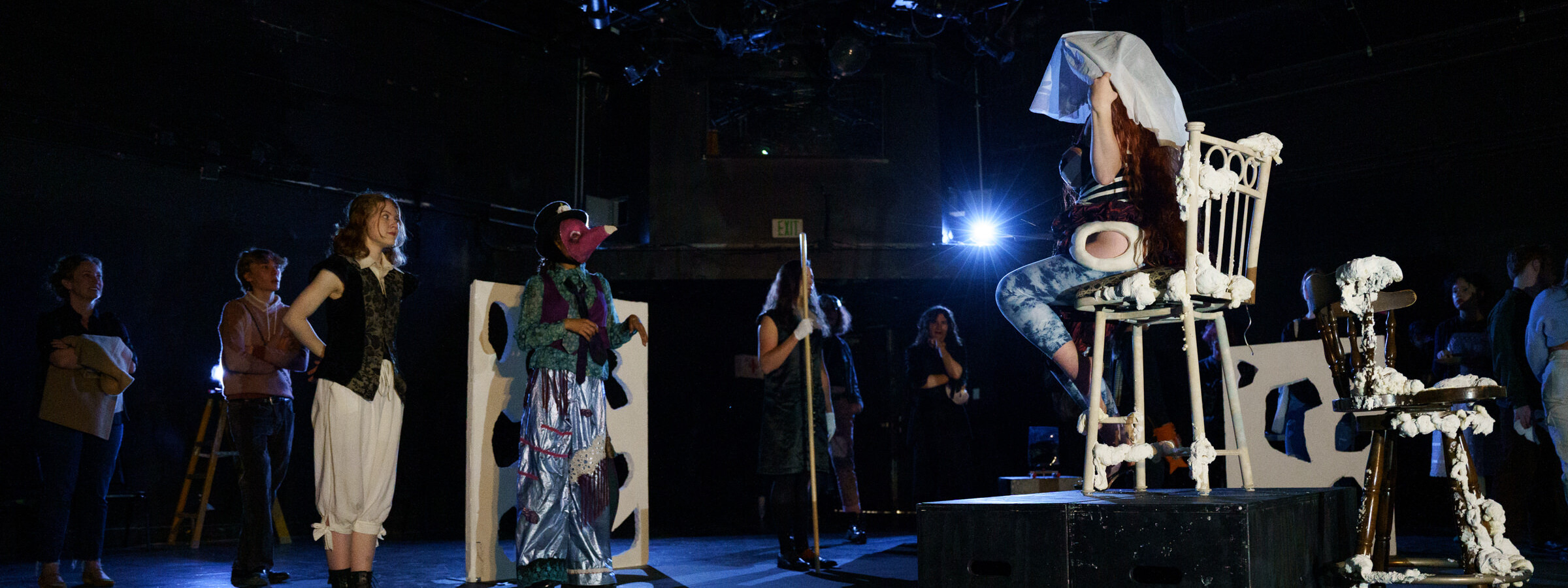 Multiple students in costume on a dark stage face another student, seated in a raised chair.