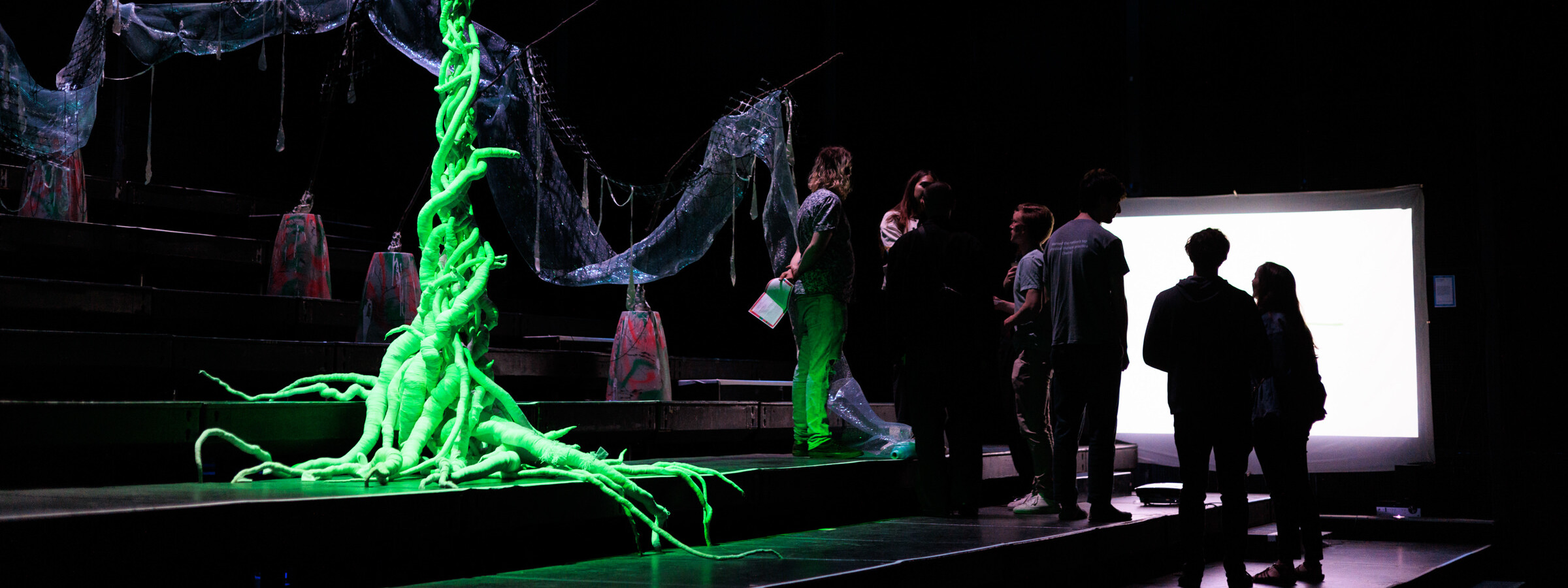 A dark theater space, with a group of people silhouetted against a bright screen. They stand near a neon green tree-like sculpture.