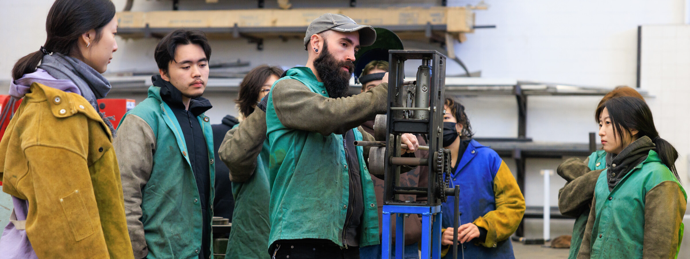 a group of people in work coats gather around a piece of machinery in CalArts Super Shop