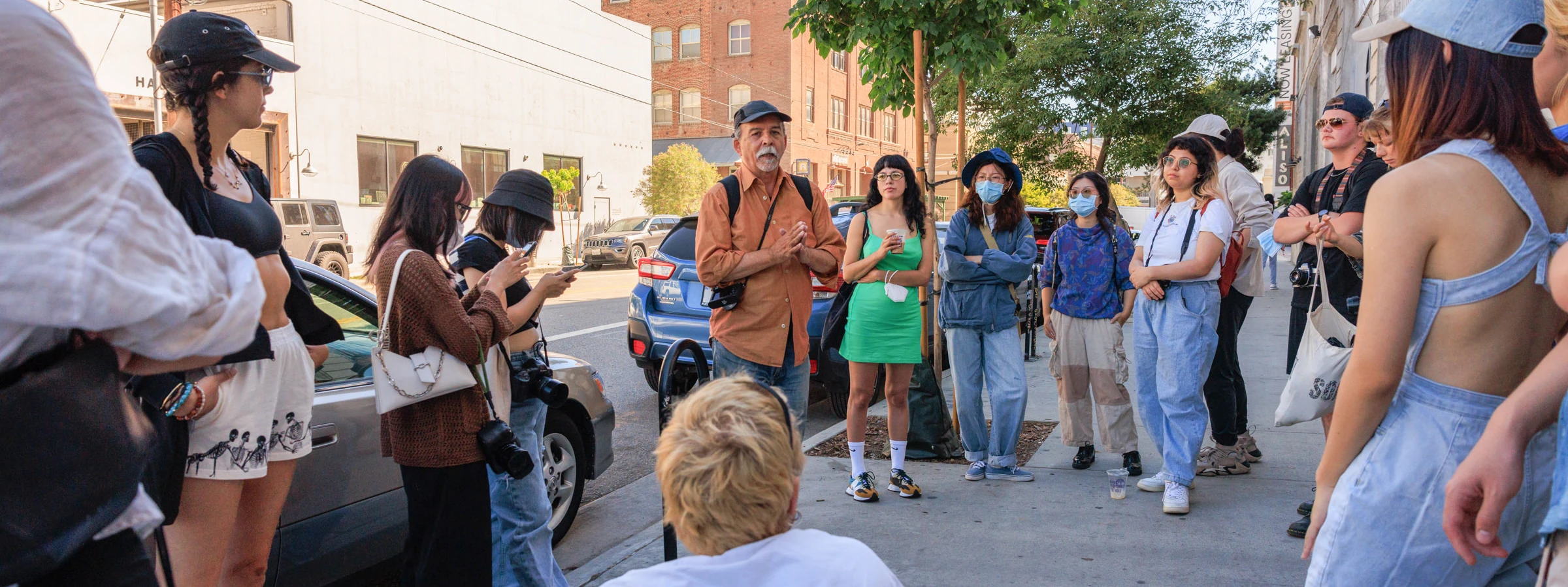 A CalArts professor in a baseball cap speaks to a large group of students assembled on a city sidewalk