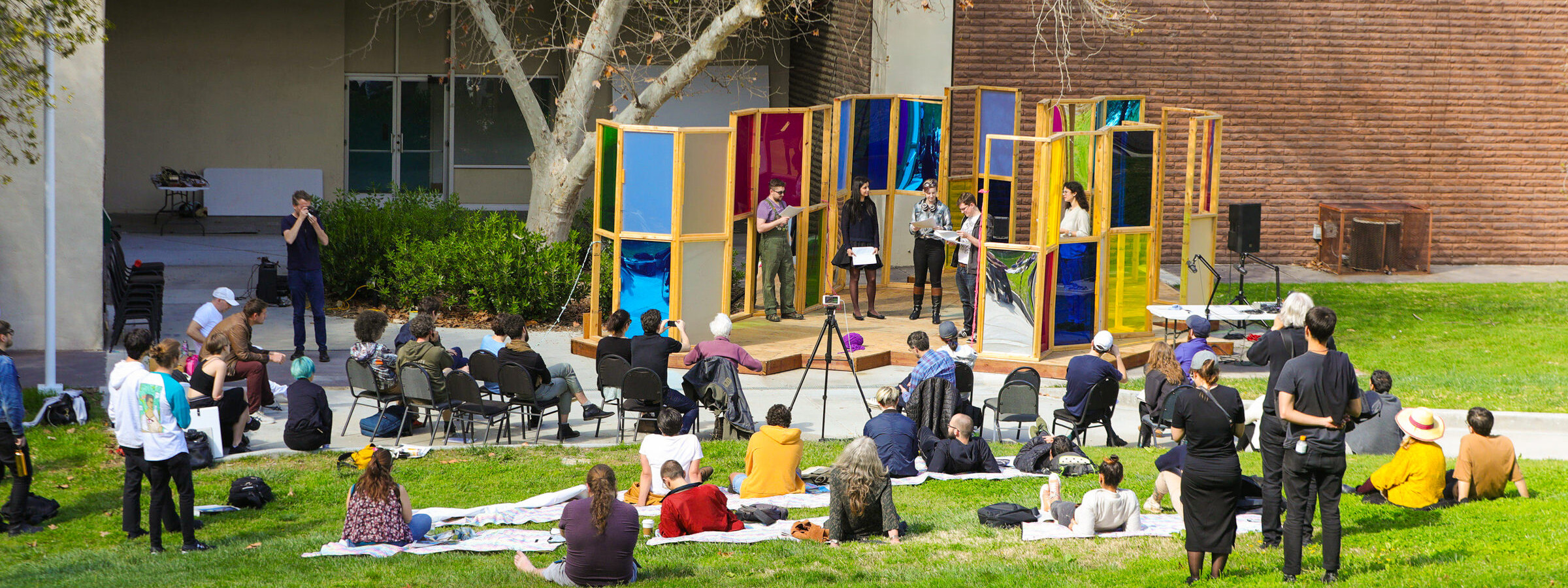 outdoor theater performance with audience members casually sitting on the lawn in front of the set outside of a school building