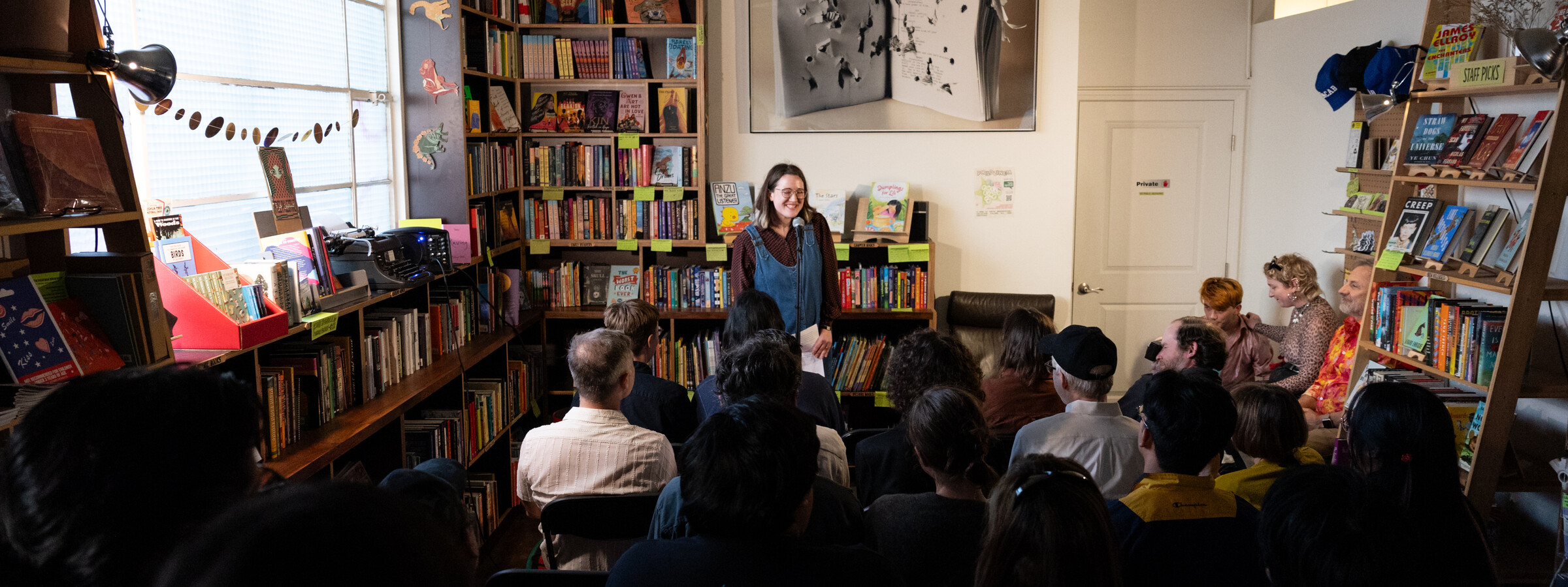 A person at a microphone addresses a small group of people seated in a cozy room lined with bookshelves. 