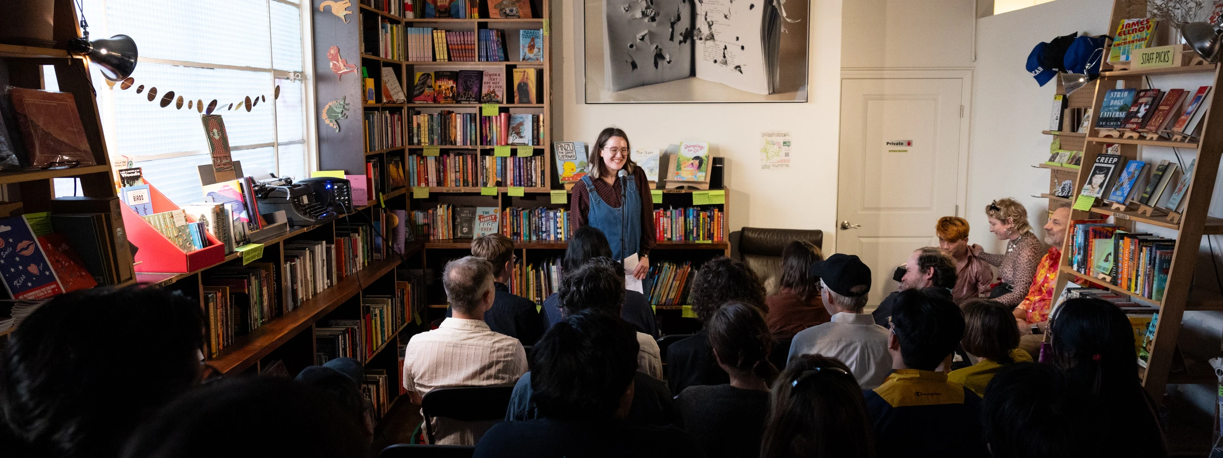 A person at a microphone addresses a small group of people seated in a cozy room lined with bookshelves. 