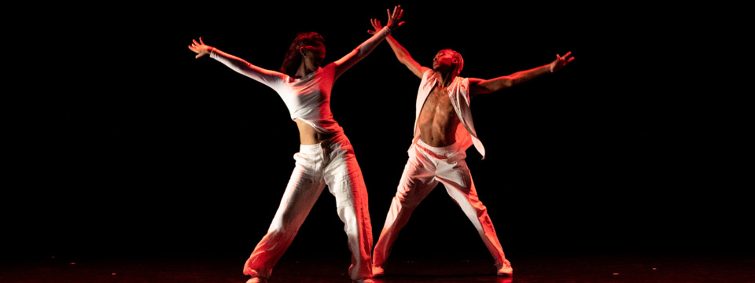 Two dancers stand on a dark stage, turned slightly toward each other, with their arms and legs spread out. 