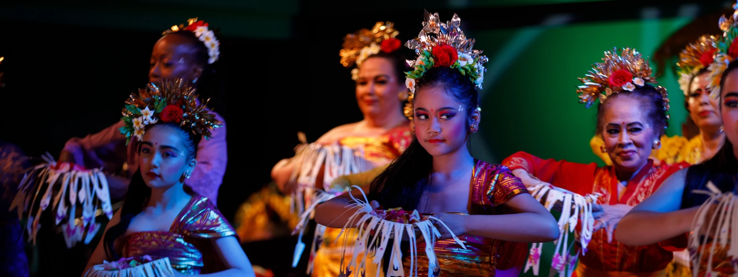 A row of dancers on stage, dressed in elaborate, colorful traditional costumes performing a Balinese dance.