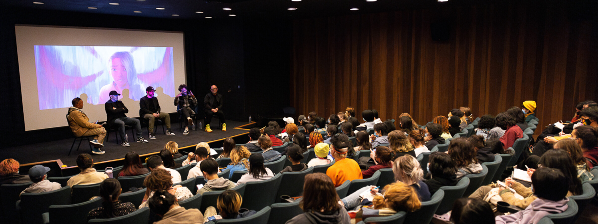 A small theater with every seat filled with a panel of speakers on stage