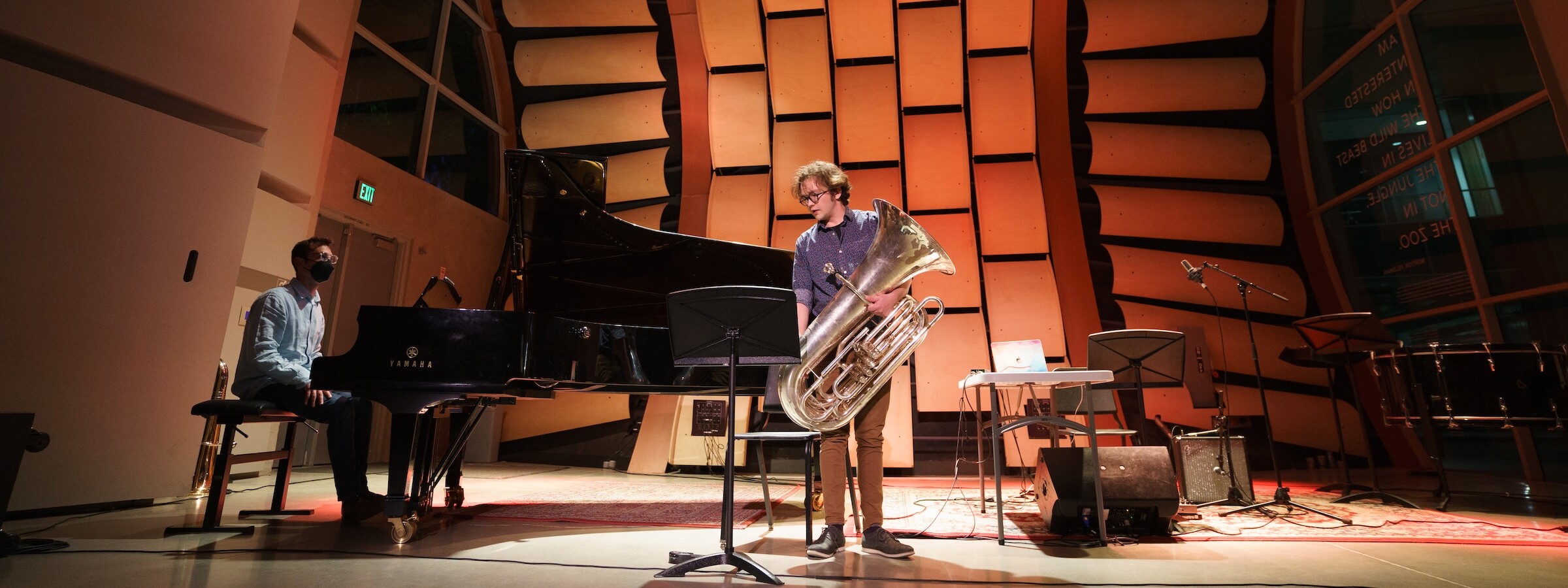 A musician plays a grand piano and another holds a trumpet on a large, modernistic, mostly empty stage