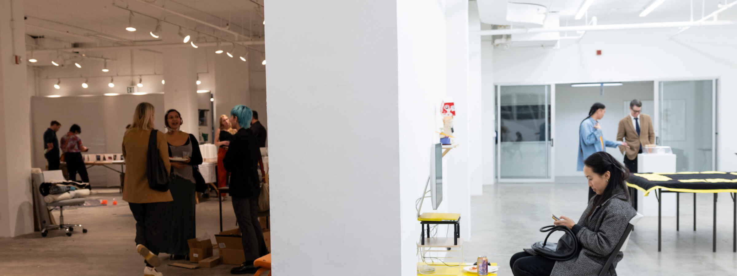 Reef gallery during an opening, showing students mingling on one side in dim light, and around the corner a student sits to work in bright light. 