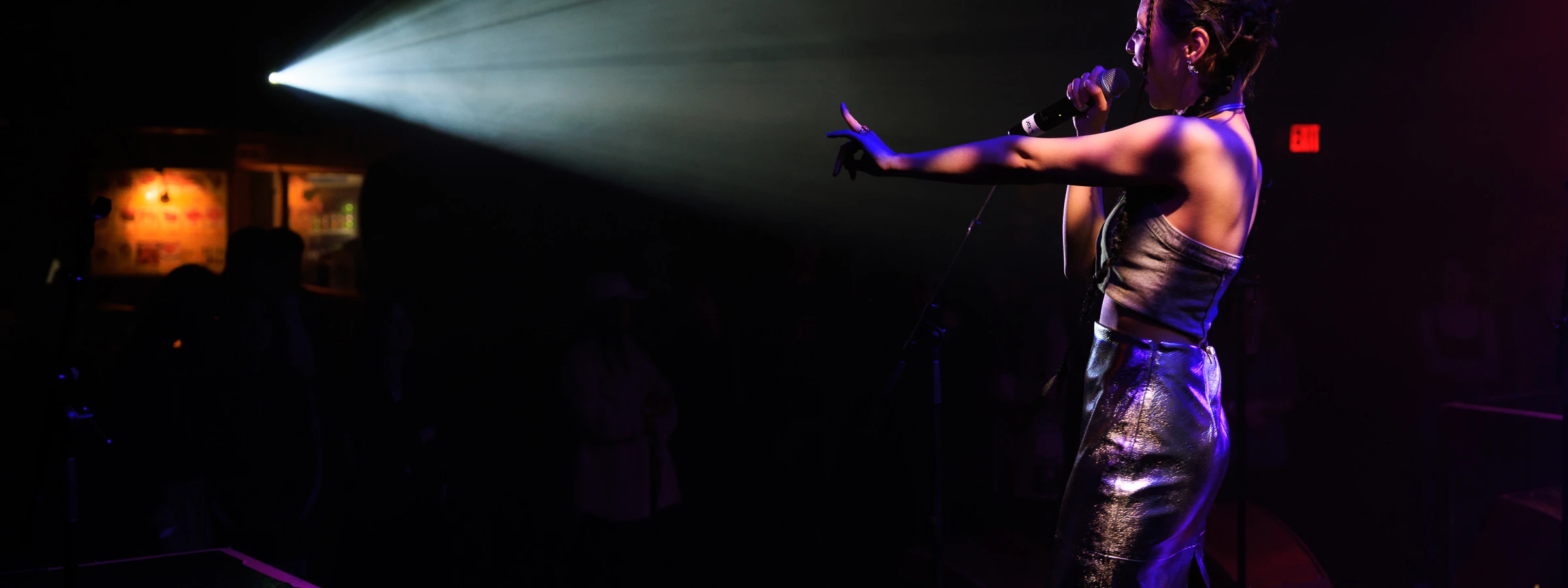 A woman sings into a microphone while holding one arm outstretched. Light from a projector streams toward her in a darkened space.