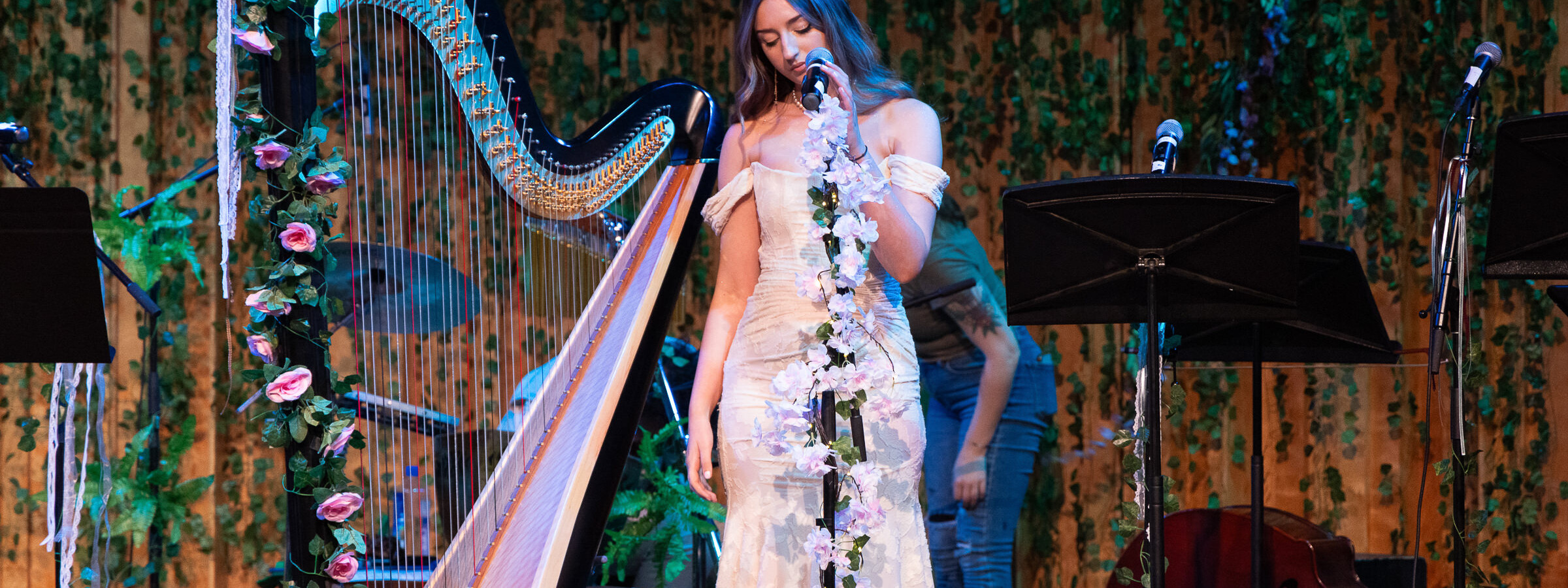 A young woman in a white dress stands at a microphone. Next to her is a large harp wrapped in flowers.