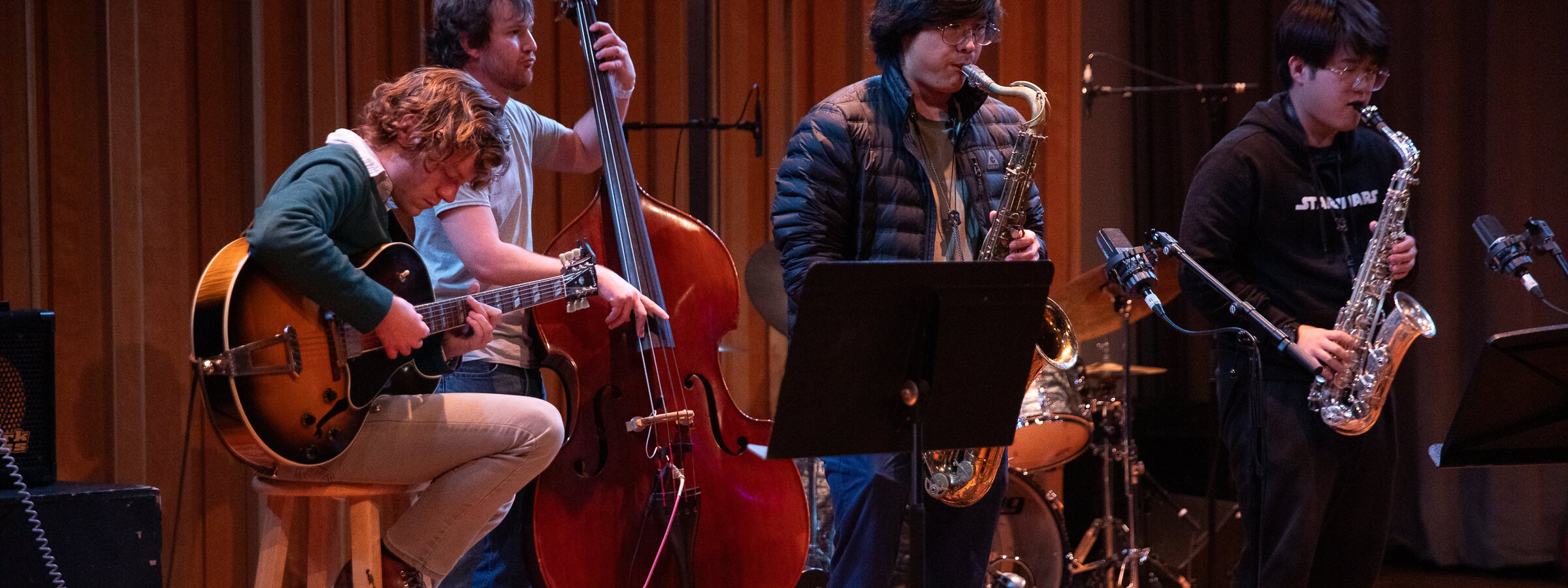 A group of casually dressed young men plays jazz instruments, including a guitar, stand-up bass, two saxophones, and drum set.