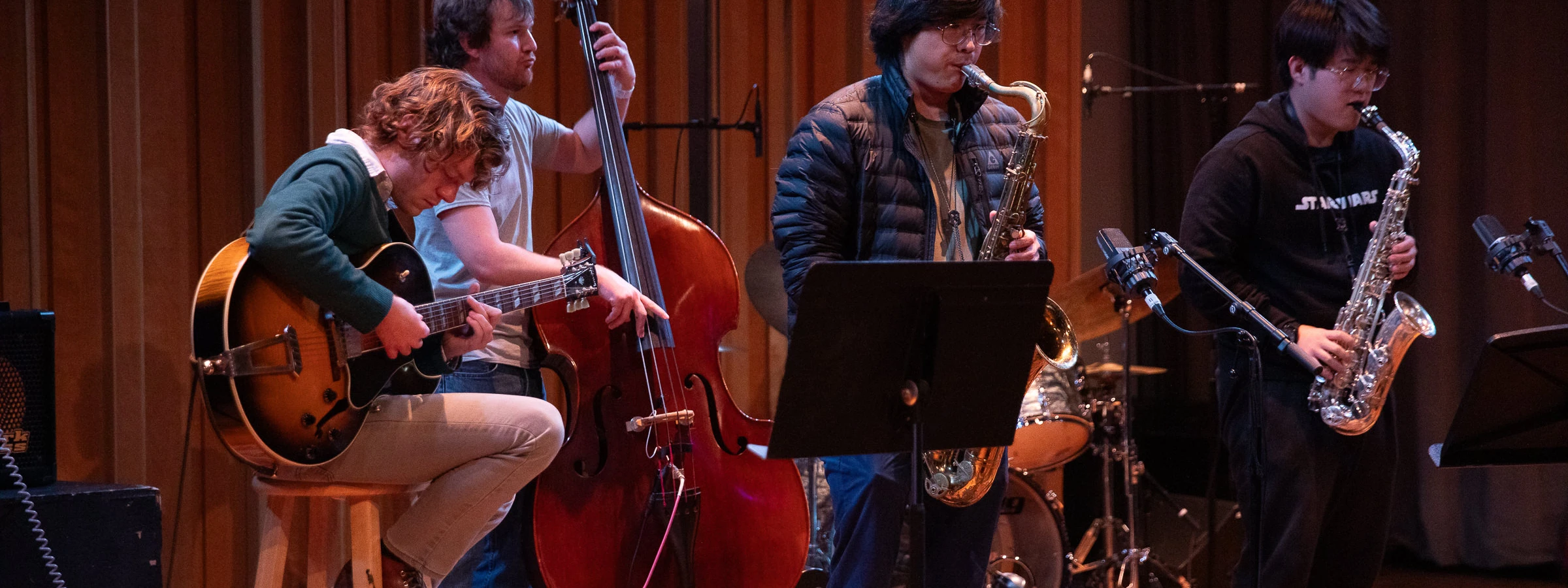 A group of casually dressed young men plays jazz instruments, including a guitar, stand-up bass, two saxophones, and drum set.
