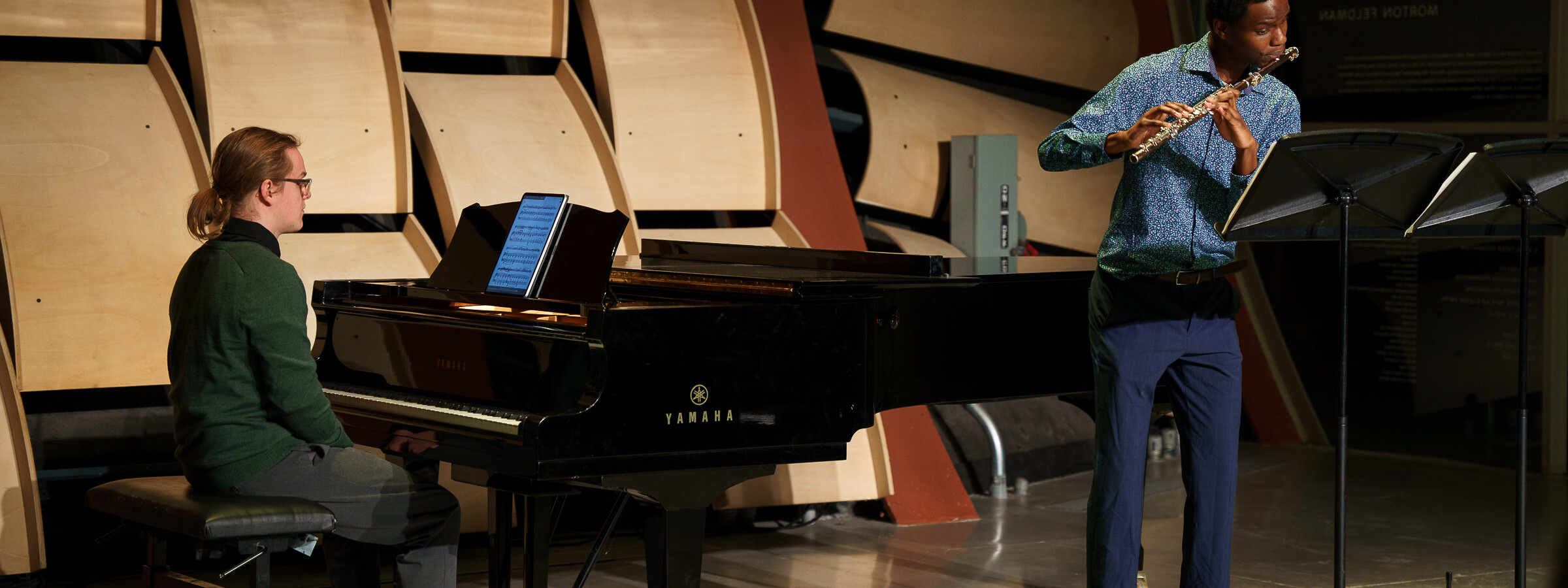 A man sits at a black, baby grand piano on a modernistic stage while another man stands at a microphone playing a flute.