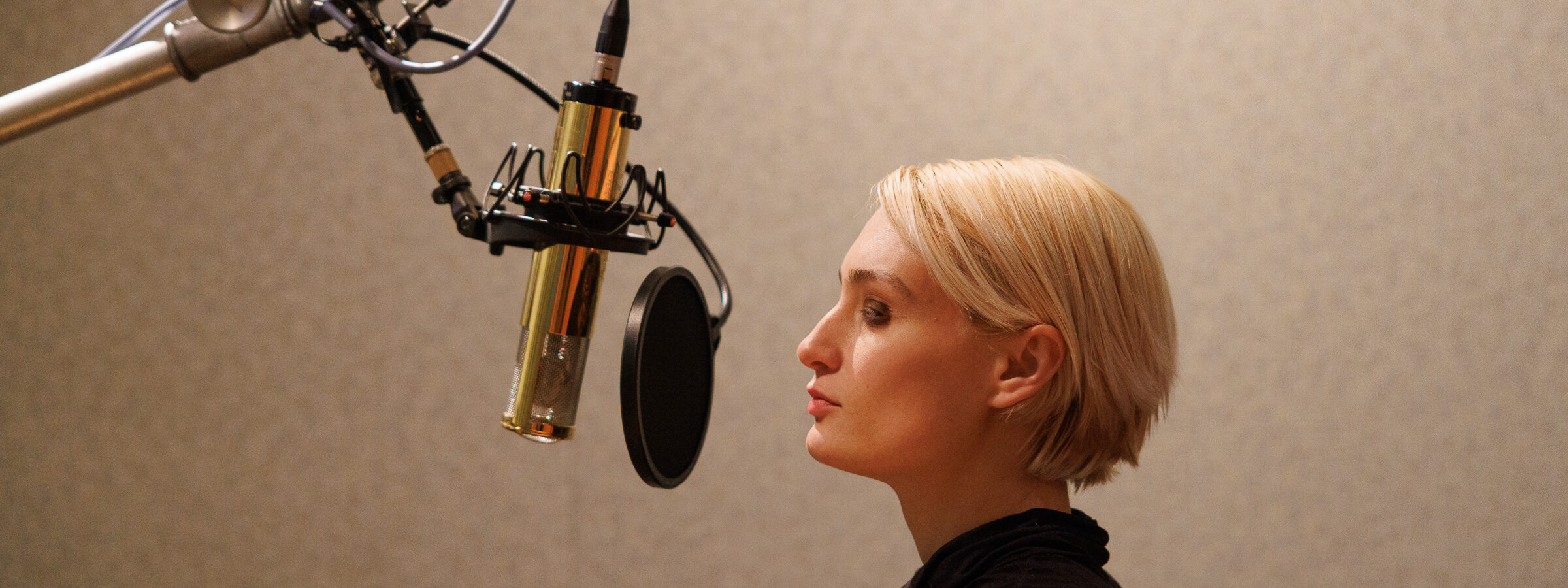 A young woman with short blonde hair is seen in profile from the shoulders up, standing at a microphone with her eyes closed.