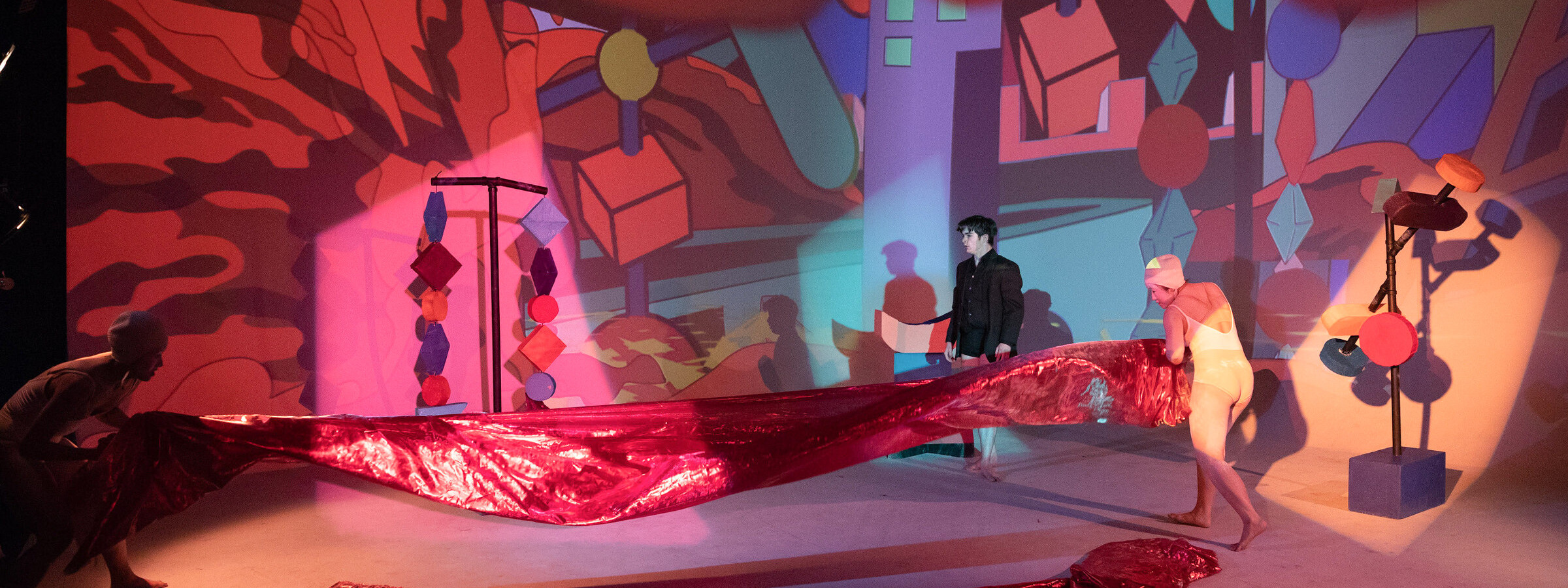 Performers stand on a brightly colored stage, holding a large banner. The stage is covered in geometric shapes. 