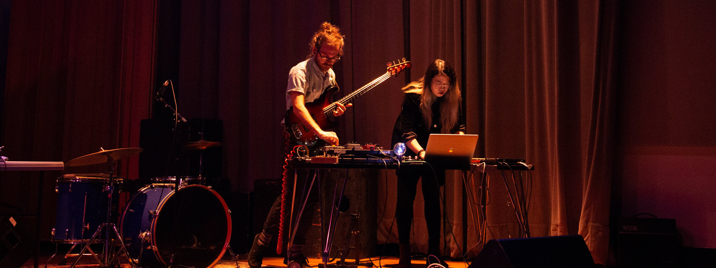 Two individuals performing on stage with musical and electronic equipment. One is playing an electric bass guitar, and the other is operating a laptop.