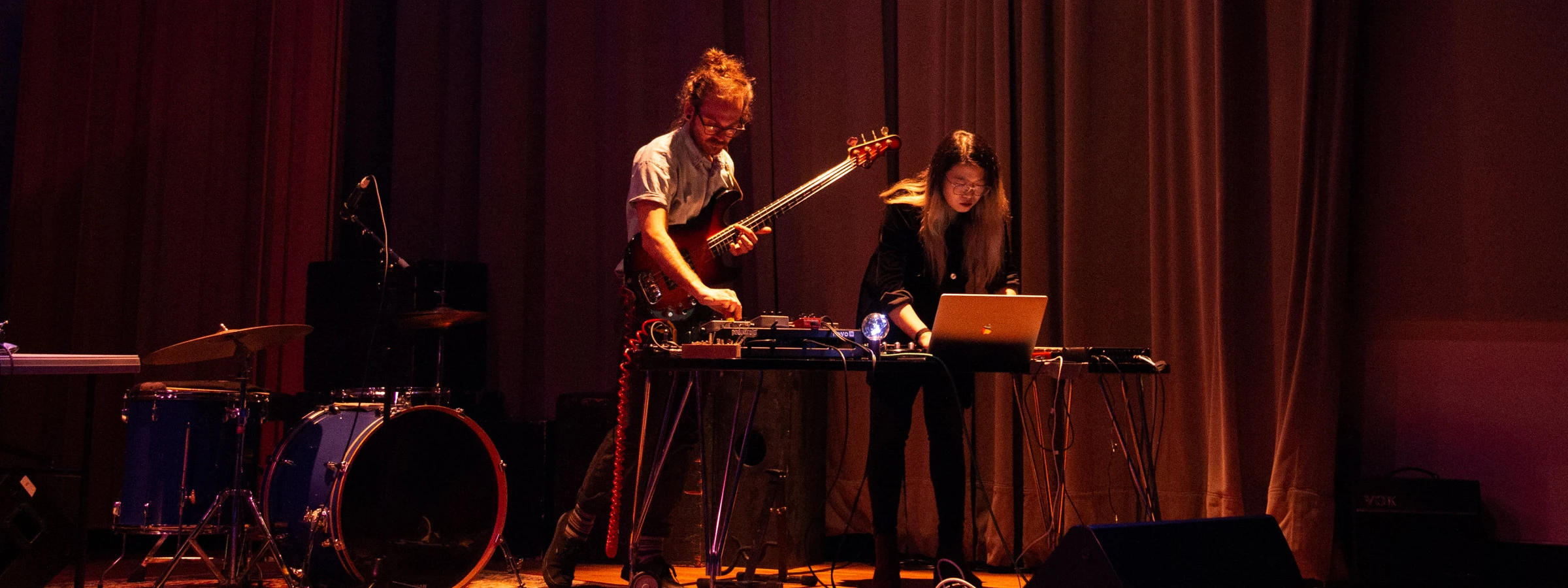 Two individuals performing on stage with musical and electronic equipment. One is playing an electric bass guitar, and the other is operating a laptop.