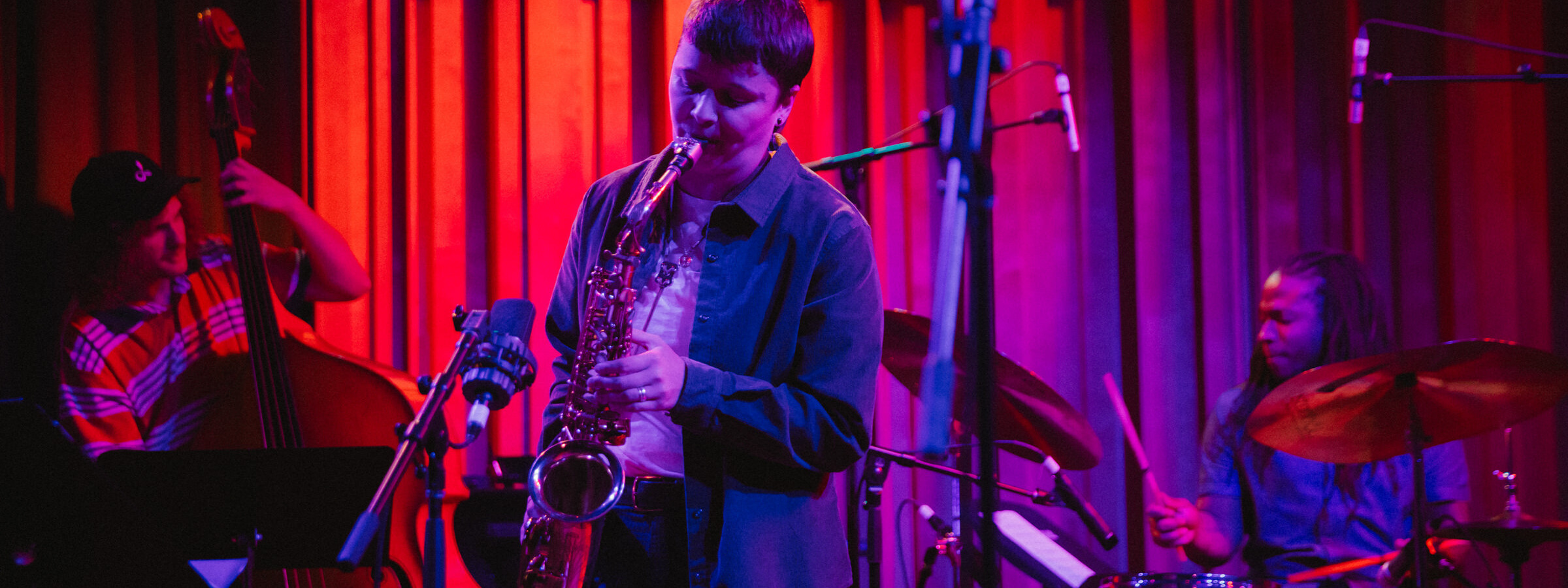 A person plays a saxophone on a small stage with moody pink and purple light. Musicians playing a standup bass and a drum set are in the background.