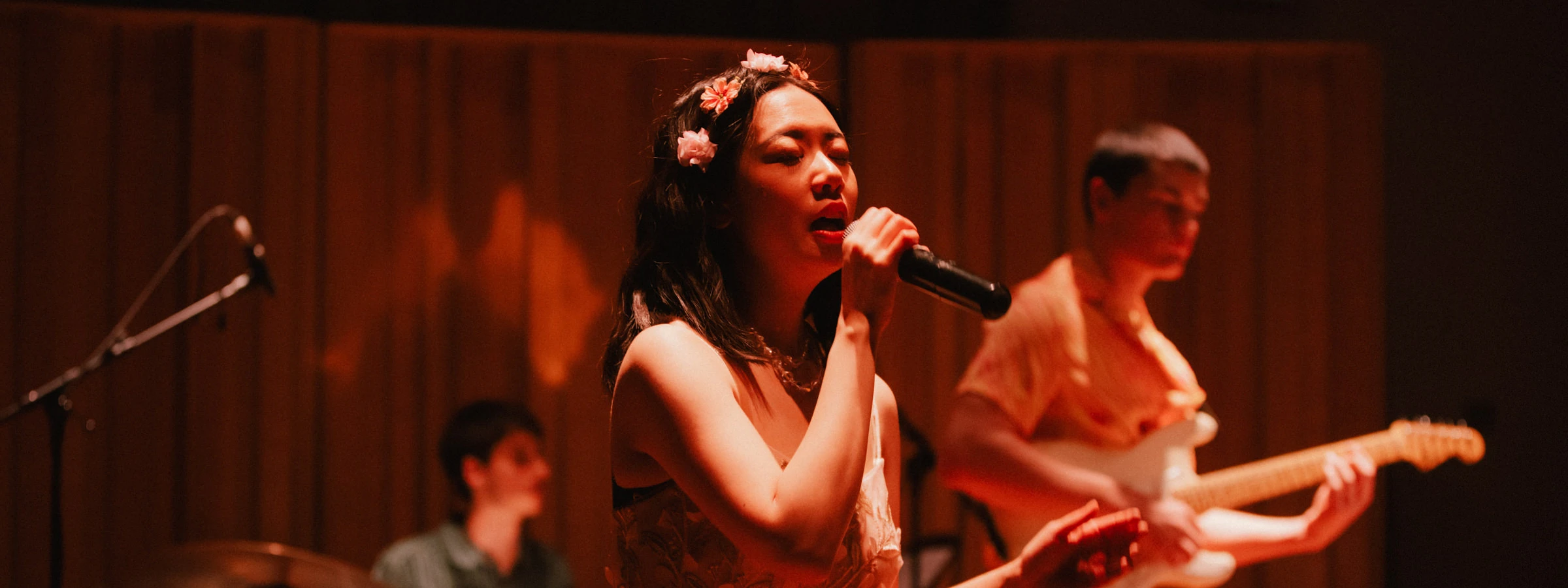 A woman in a flower crown sings intensely into a microphone, bathed in pink light. 