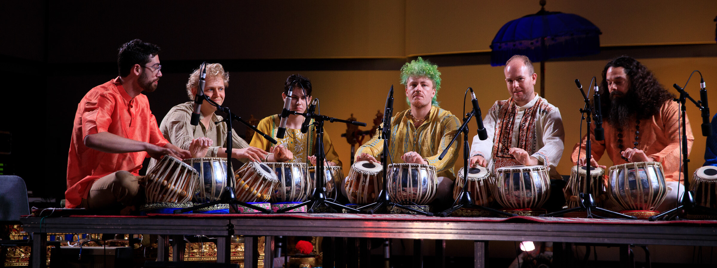 A group of tabla drum players gathers on a stage, with microphones positioned at their drums. 