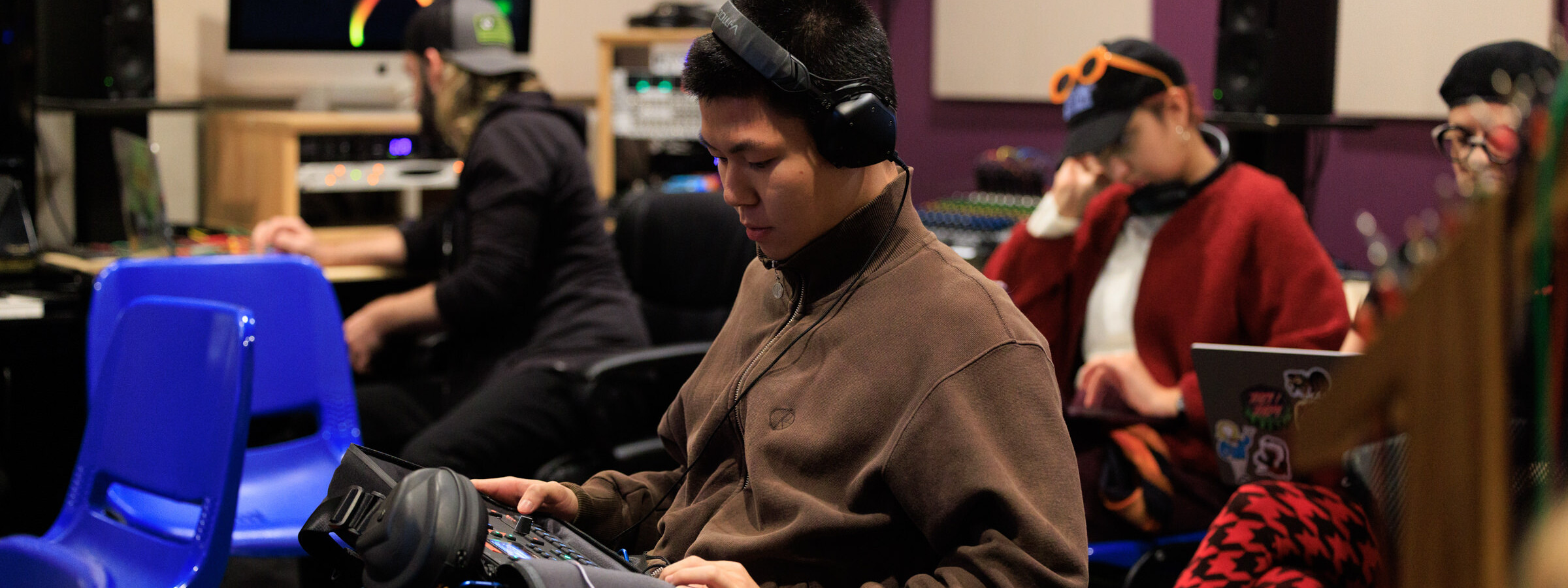 A person sits in a college classroom with other students surrounded by tech equipment. They are holding a small sound mixing board in their lap.