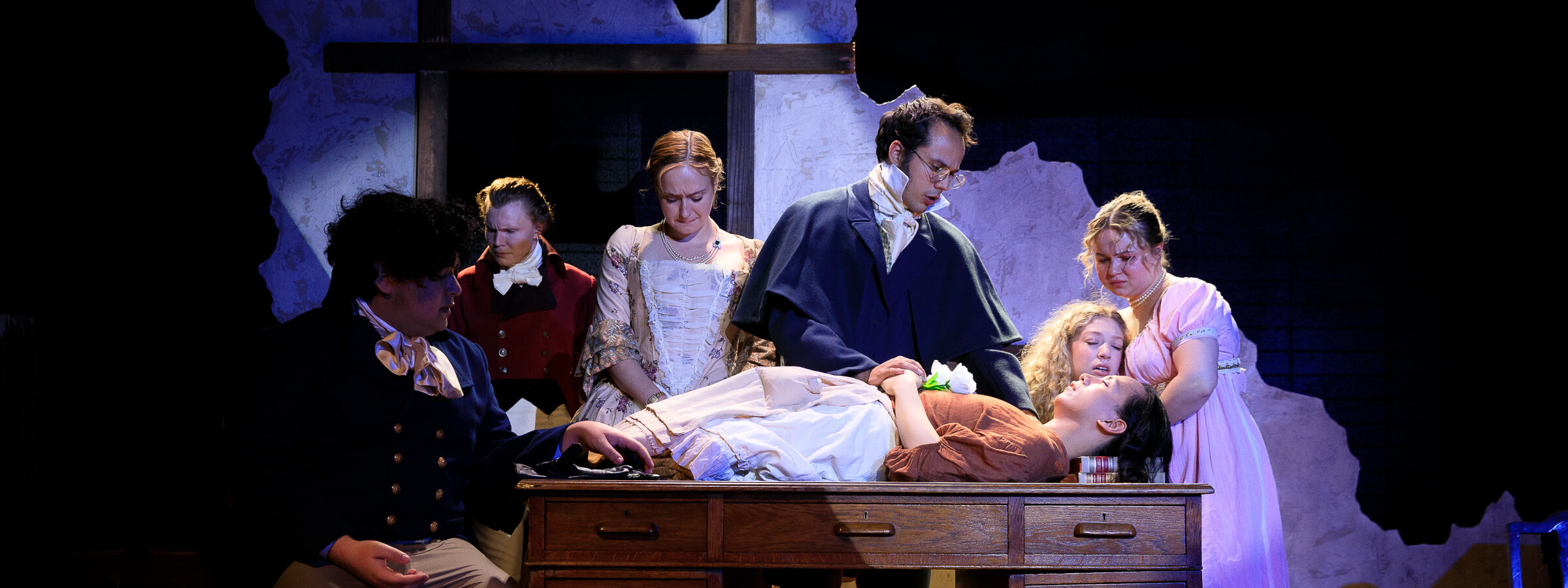 Play on a stage depicting a recently deceased person lying on a wooden desk as others surround her and mourn her body. All are dressed in colonial-era costume.