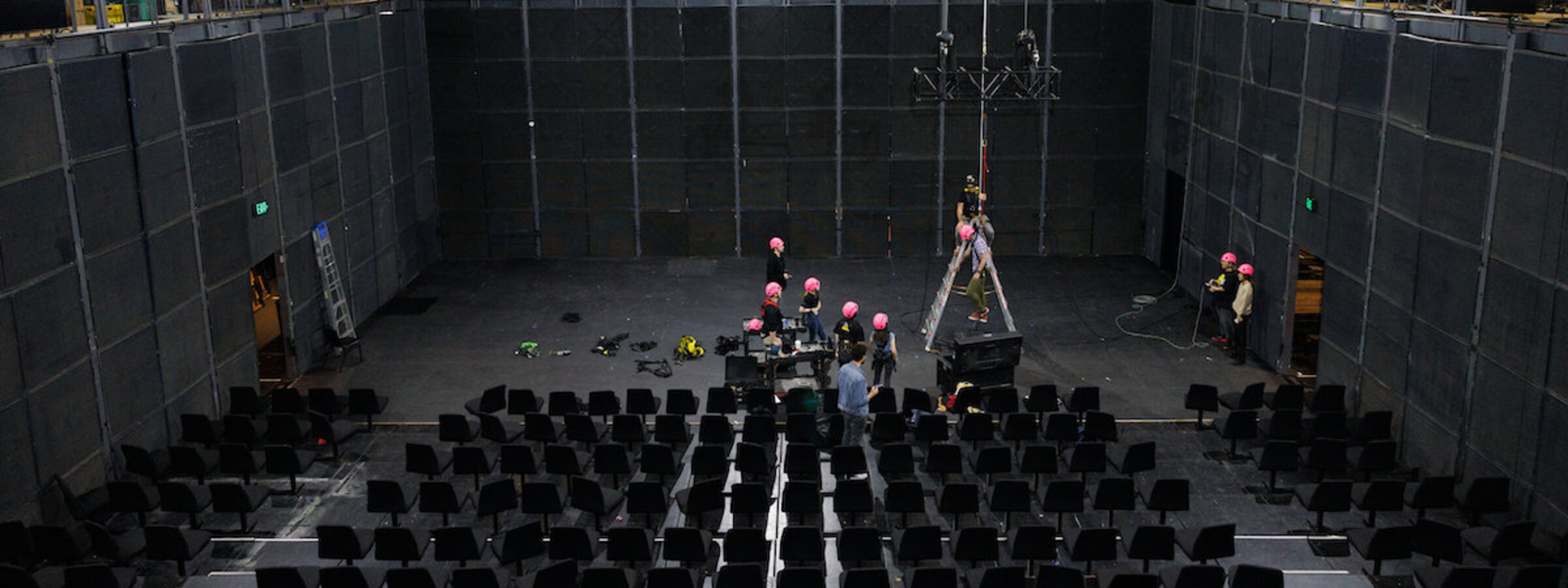 A group of students in pink helmets practice harness safety in a giant theater at CalArts