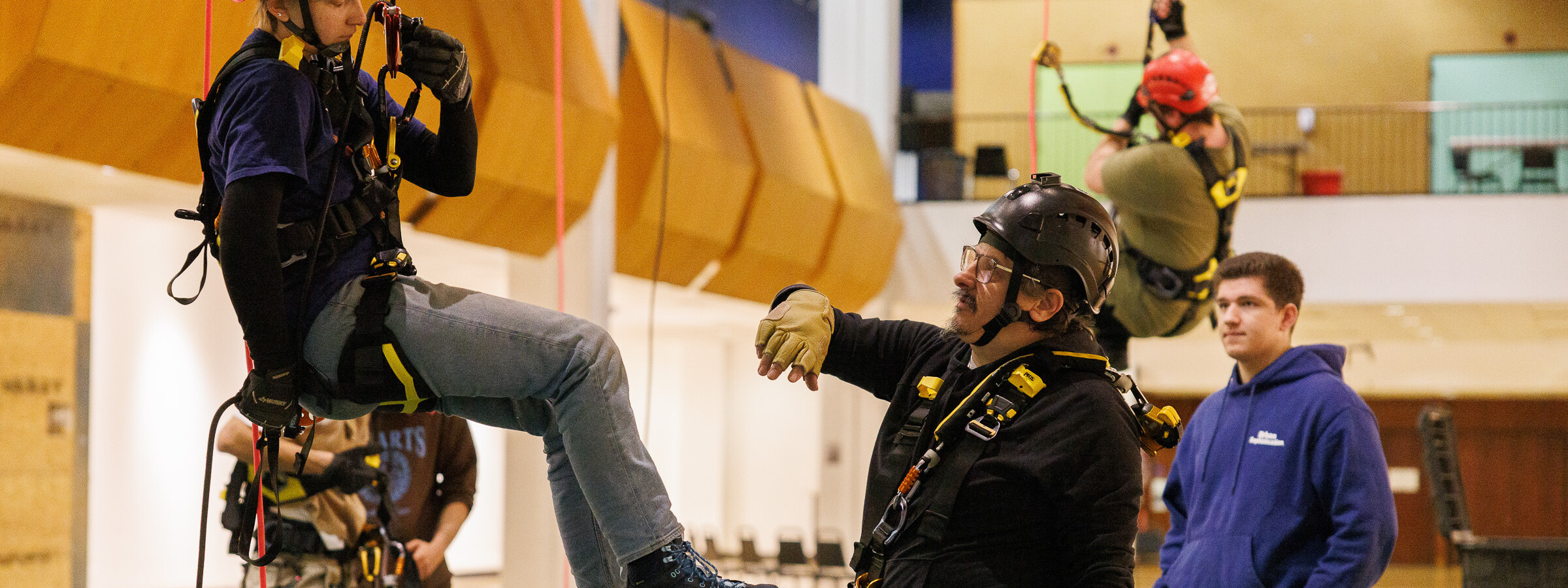 Person wearing a hard hat and strapped into a harness hangs in the air in an indoor theatrical space as another person, standing on the floor, directs them. 